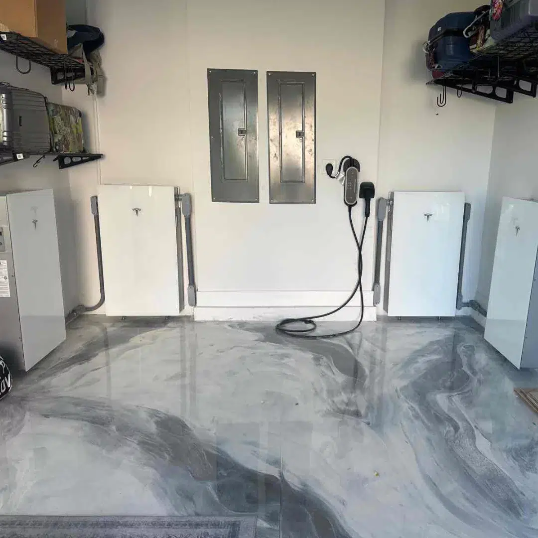 A modern garage interior with a clean, polished marble floor. Mounted on the wall are four Tesla Powerwall battery units, two electrical panels, and a Tesla EV charger. Storage shelves with tools and equipment are visible on both sides.