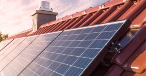 Solar panels with multiple cells placed on a roof with metal brown shingles next to a chimney on a sunny day.