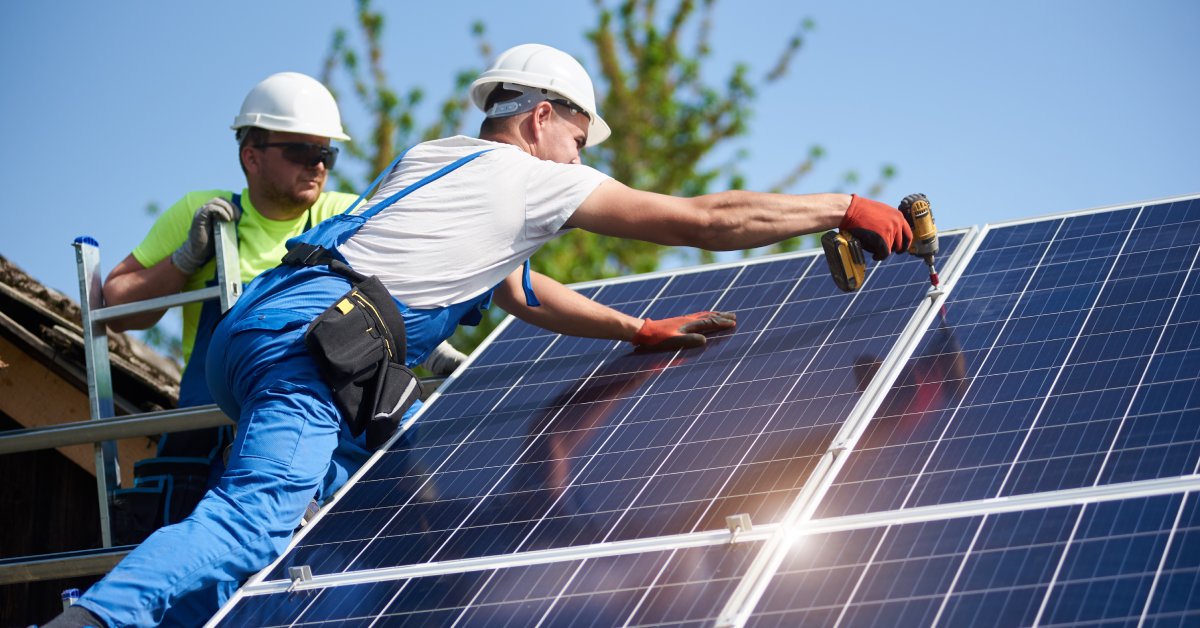Two workers wearing helmets and protective gloves standing on top of a roof installing multiple large solar panels.