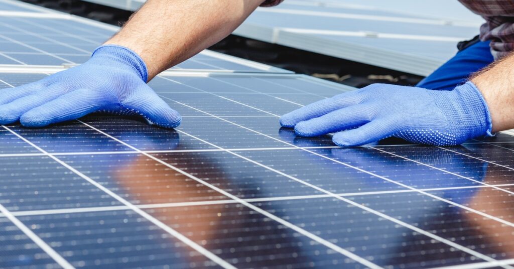Tesla Powerwall Explained: A Smart Backup Power Solution 2 A close-up of two hands wearing blue protective gloves touching a large solar panel with multiple cell rows on a roof.