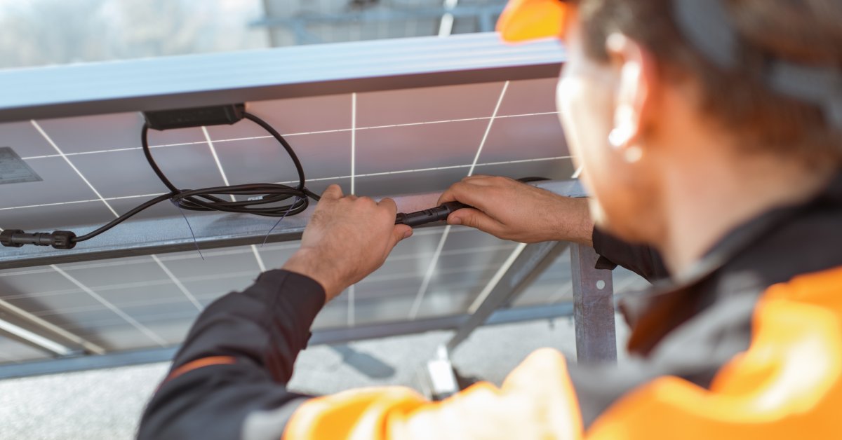 A worker wearing an orange hard hat and vest checks the connecting cables on the back of a large solar panel.