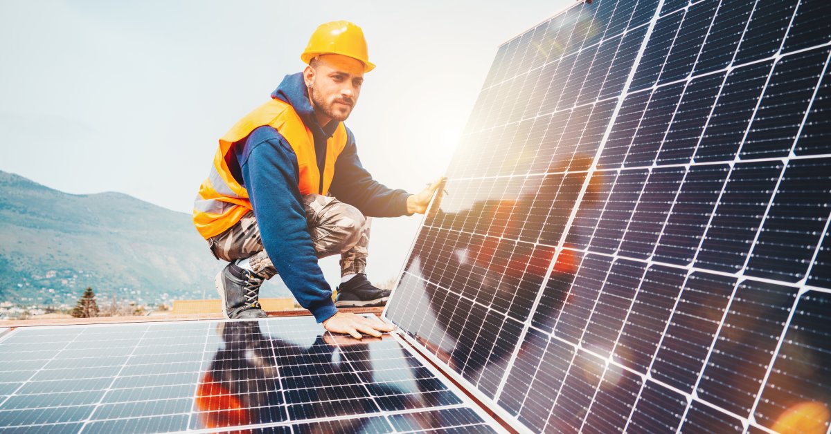 A worker wearing an orange hard hat and safety vest squats while installing a large solar panel on a roof.