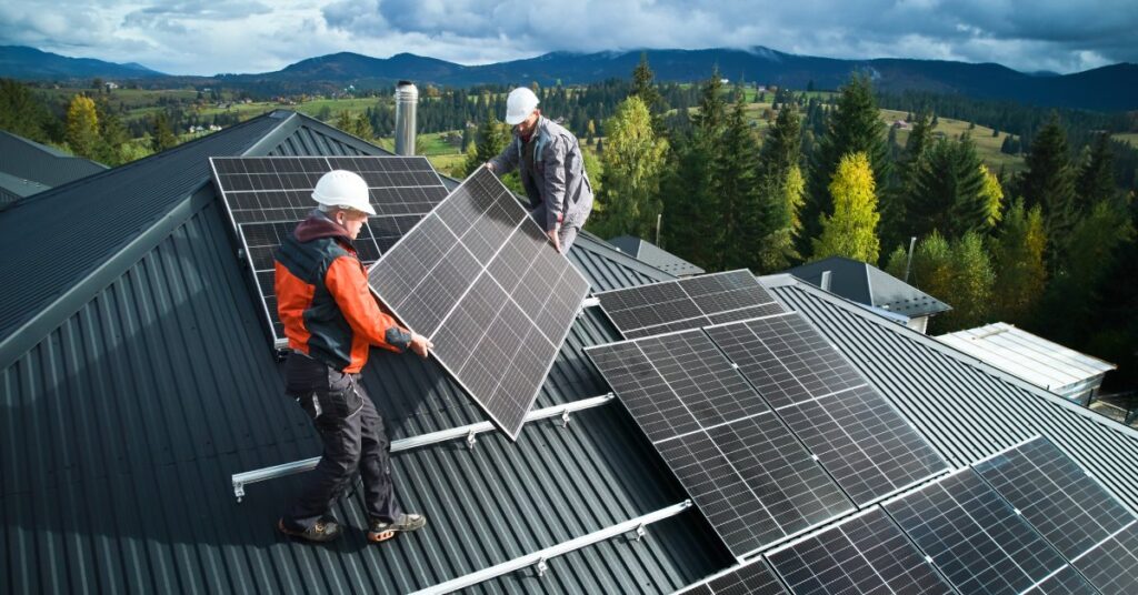 Two workers wearing white hard hats install a large solar panel on the roof of a home next to other panels.