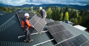 Two workers wearing white hard hats install a large solar panel on the roof of a home next to other panels.