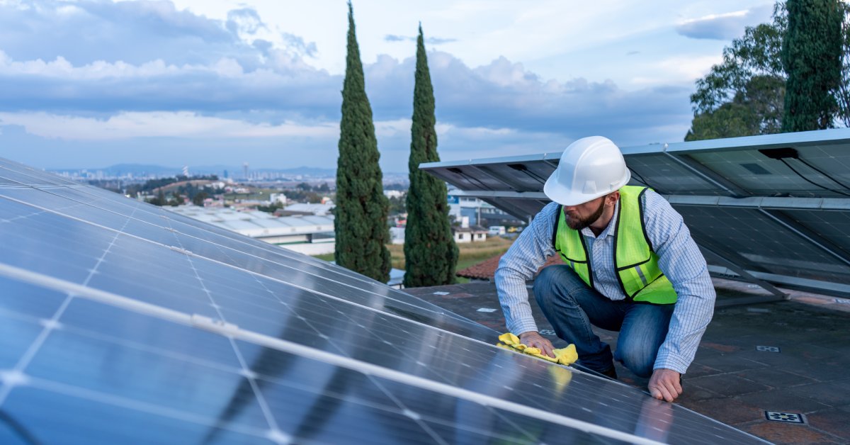 A worker wearing a vest and white hard hat uses a yellow cloth to clean a large row of solar panels on a roof.