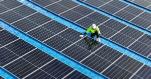 A worker wearing a bright yellow jacket and a white hard hat kneels next to a row of solar panels on a roof.