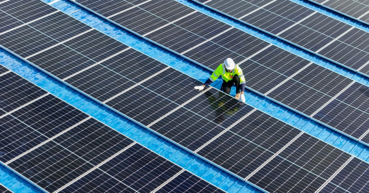 Learn 4 A worker wearing a bright yellow jacket and a white hard hat kneels next to a row of solar panels on a roof.