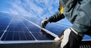 Close-up of a man wearing safety gloves and a jacket securing a large solar panel next to other panels on a roof.