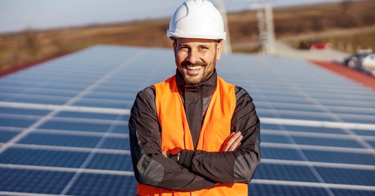 Worker wearing a white helmet and a safety vest smiling while standing in front of multiple solar panels on a roof.