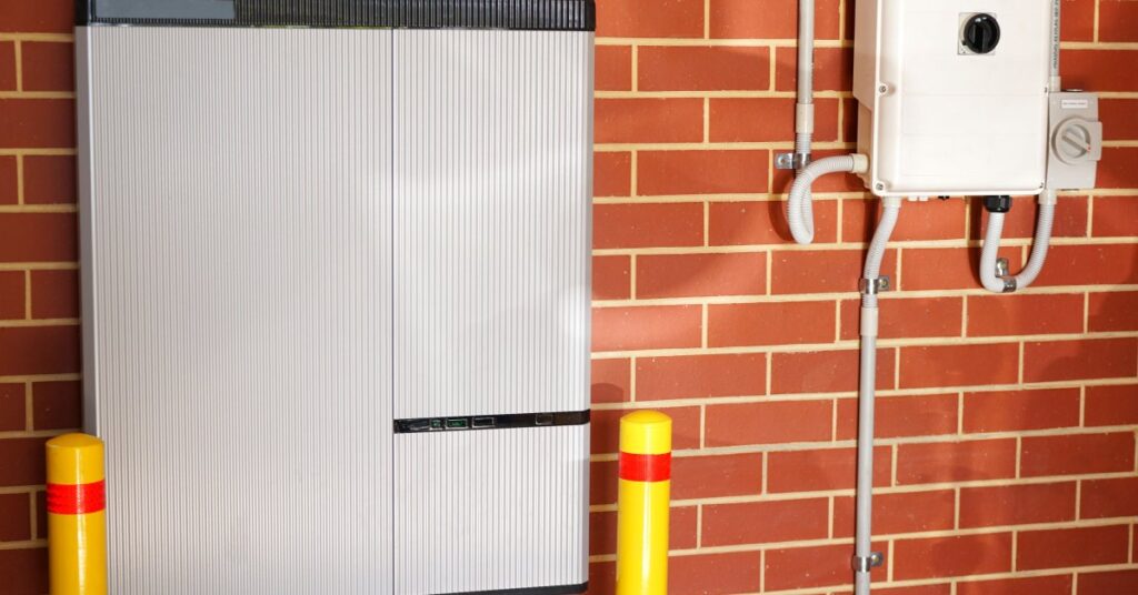 A large white home battery attached to a brick wall with a small transformer on the side attached to white tubes.