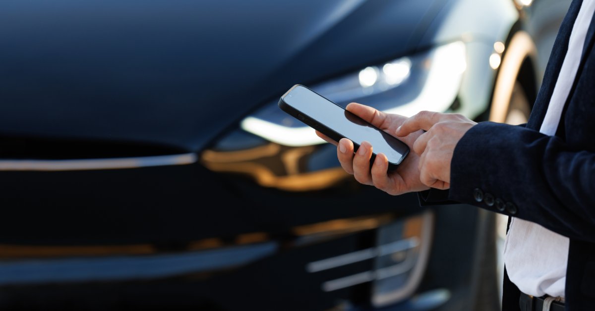 The arms of a man wearing a black suit jacket. He touches his smartphone's screen as a Tesla sedan sits in the background.