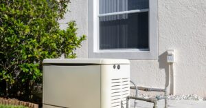 White residential power generator placed outdoors under a window, next to a bushy tree and lush green grass.