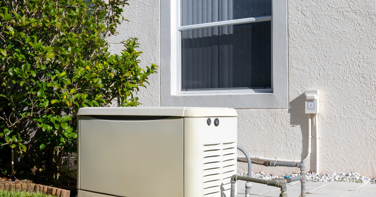 Learn 8 White residential power generator placed outdoors under a window, next to a bushy tree and lush green grass.