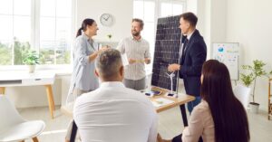 A group of people gathered in a meeting room, talking together. One man is holding a small solar panel on top of a desk.