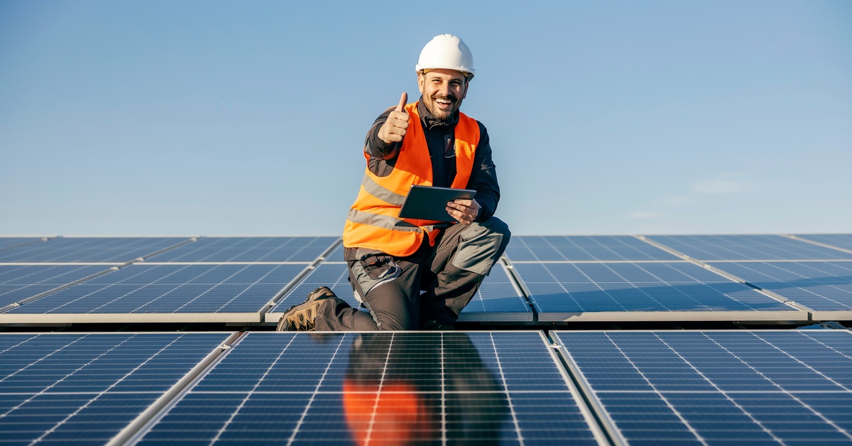 Top Questions To Ask Your Solar Power Solutions Company 2 A worker in a safety vest and white hard hat kneeling next to solar panels on a roof while giving a thumbs up.