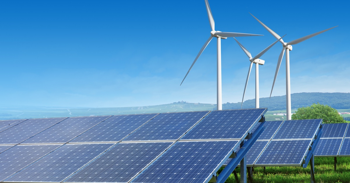 Multiple rows of solar panels are on a structure above lush grass. Three big wind turbines are in the distance.