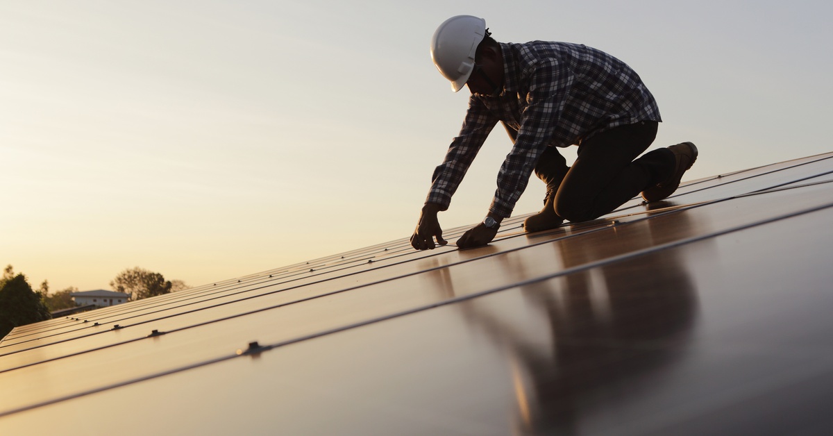 A worker wearing a plaid shirt and a white helmet is kneeling on top of a solar panel and touching the edge of a panel.