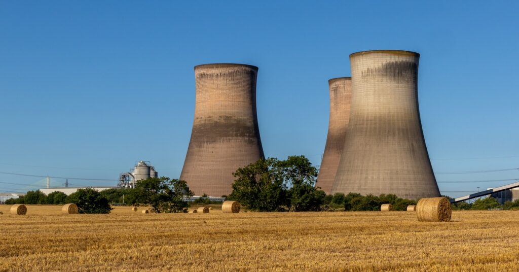 Large industrial power station with three cooling towers in the middle of a large field with multiple hay bales.