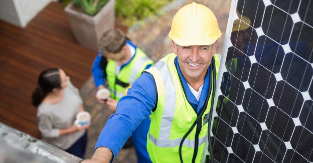 A worker wearing a yellow hard hat and safety vest climbs a ladder and carries a solar panel to the roof of a home.