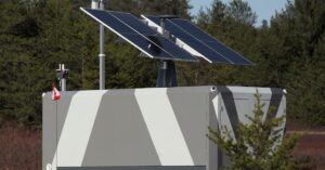 Large grey and white battery with solar panels attached to the top, placed on a plot of land surrounded by trees.