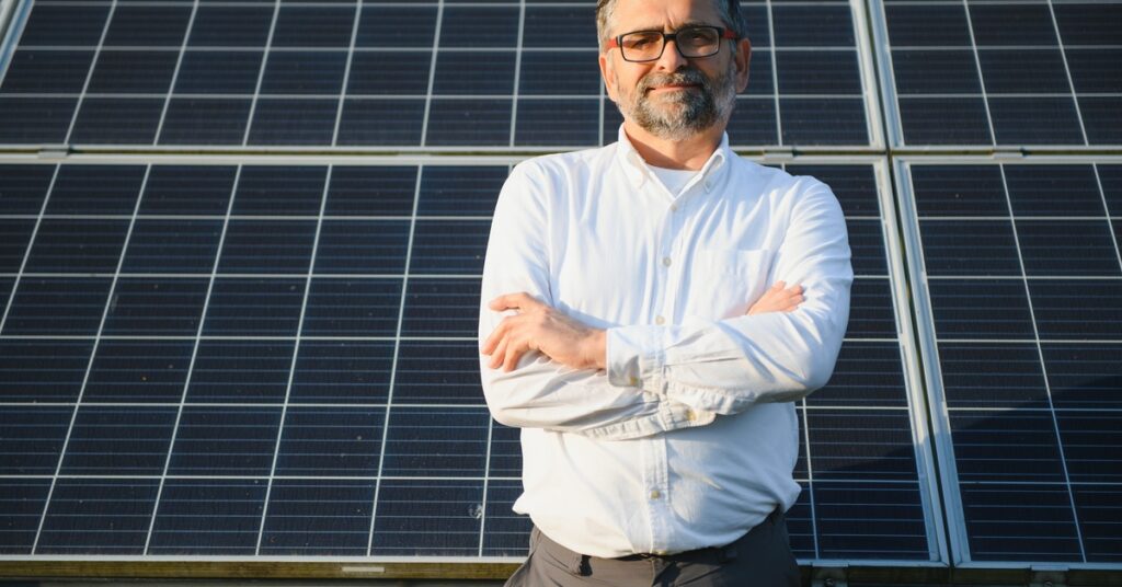Man with a beard wearing a white shirt and crossing his arms while standing in front of large solar panels.