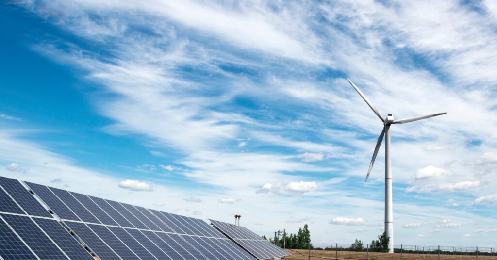 A large row of solar panels next to a wind turbine placed on an empty plot of land with some trees in the background.