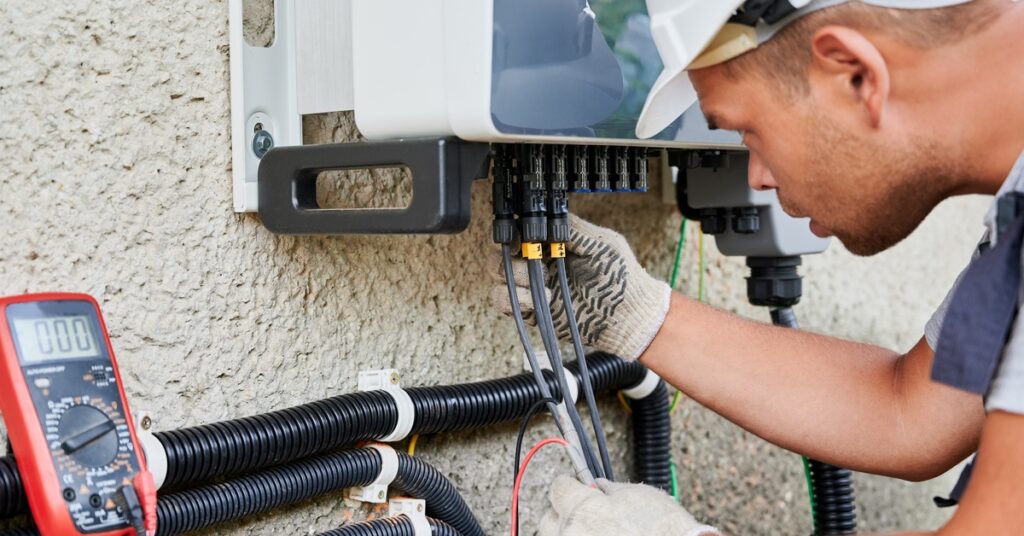 A Guide to Equipment You Need for a Solar Panel System 2 Worker wearing a white helmet inspecting the cables and connections on an electrical panel installed outdoors.