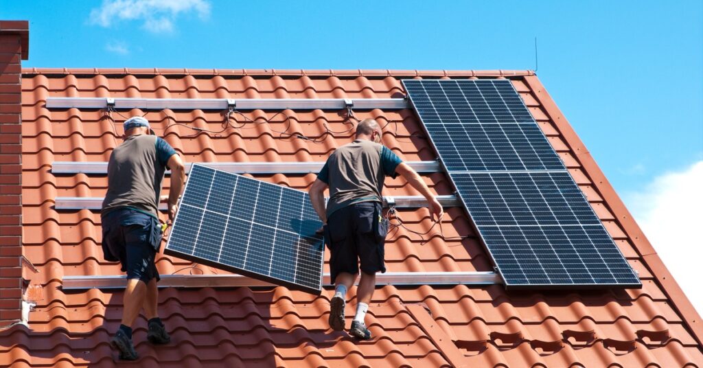 A Guide to Equipment You Need for a Solar Panel System 1 Two workers wearing brown t-shirts carrying a solar panel unit on a home roof with a white mounting system.