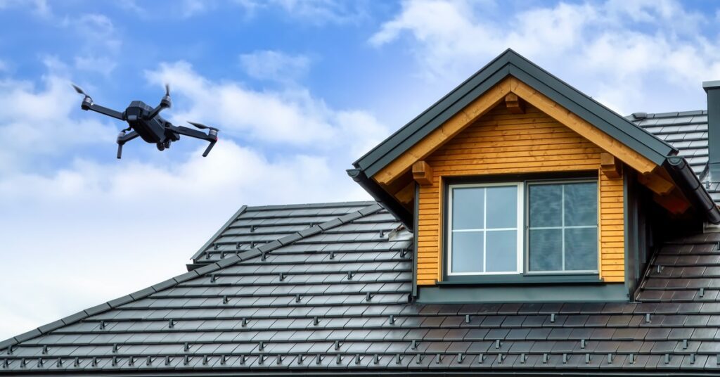 A black drone flying near the top of a two-story home with a dark roof and dormer window on the second floor.