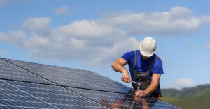 A worker wearing a blue T-shirt and white hard hat uses a wrench to tighten a solar panel installed on a roof.