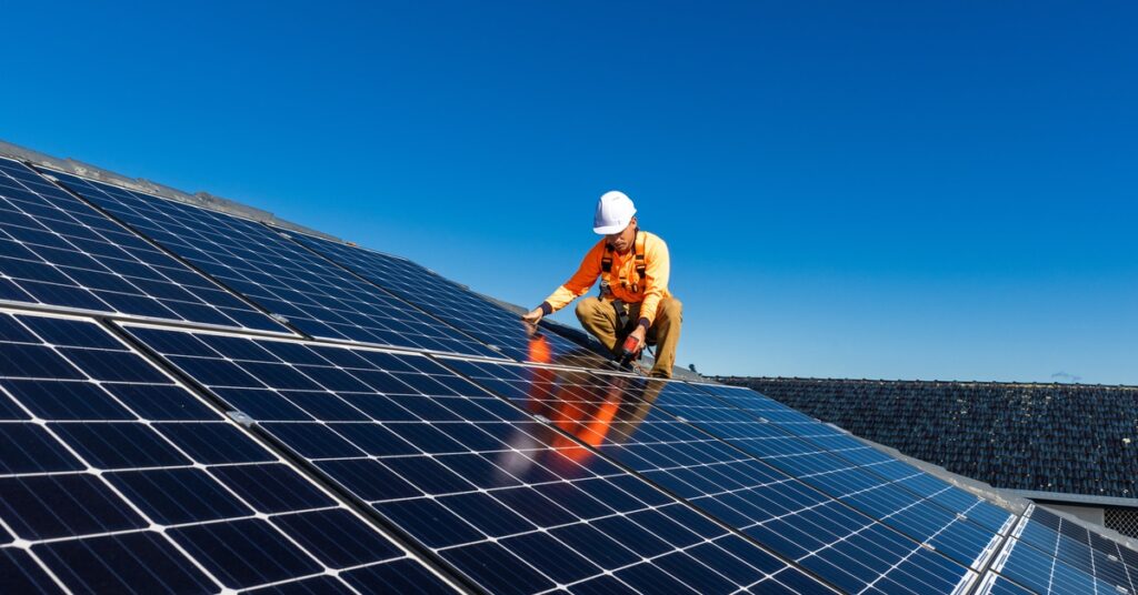 Commercial Solar Power Solutions for Warehouses 2 A man wearing a white helmet and an orange uniform touches a large solar panel on top of a roof on a sunny day.