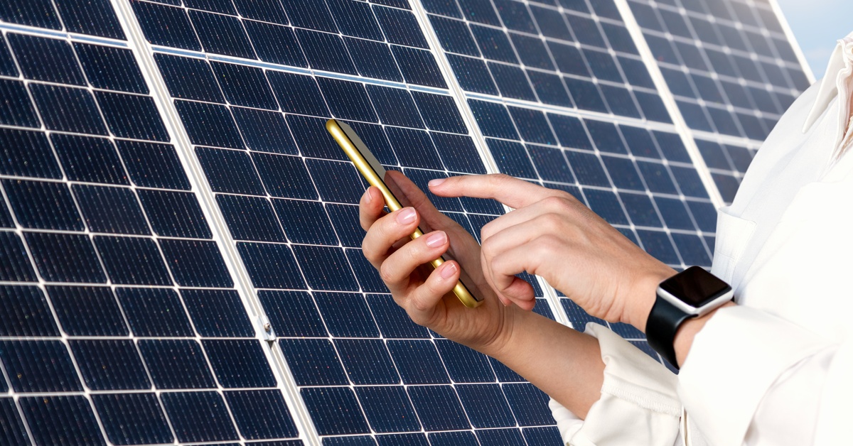 Permitting for Residential Solar Power Installation 2 A close-up of the hands of a man holding a smartphone and touching the screen in front of a large solar panel.