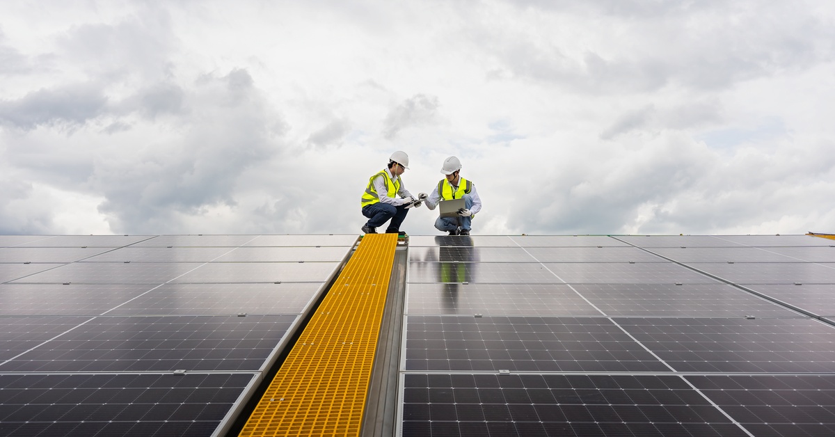 Structural Analysis for Commercial Solar Installation 1 Two workers wearing white helmets and safety vests squatting next to long rows of solar panels on a cloudy day.