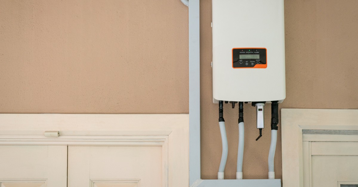 Integrating Tesla Powerwalls With Electrical Systems 1 A white solar inverter with a black frame, cable protectors, and a small digital screen, on a compacted wood wall.