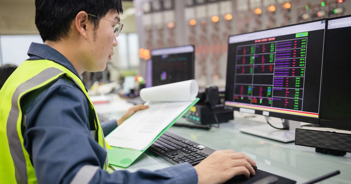 How Tesla Megapack Batteries Stabilize the Electrical Grid 1 An engineer holding a clipboard while sitting behind a desk, working on a computer monitor displaying numbers.