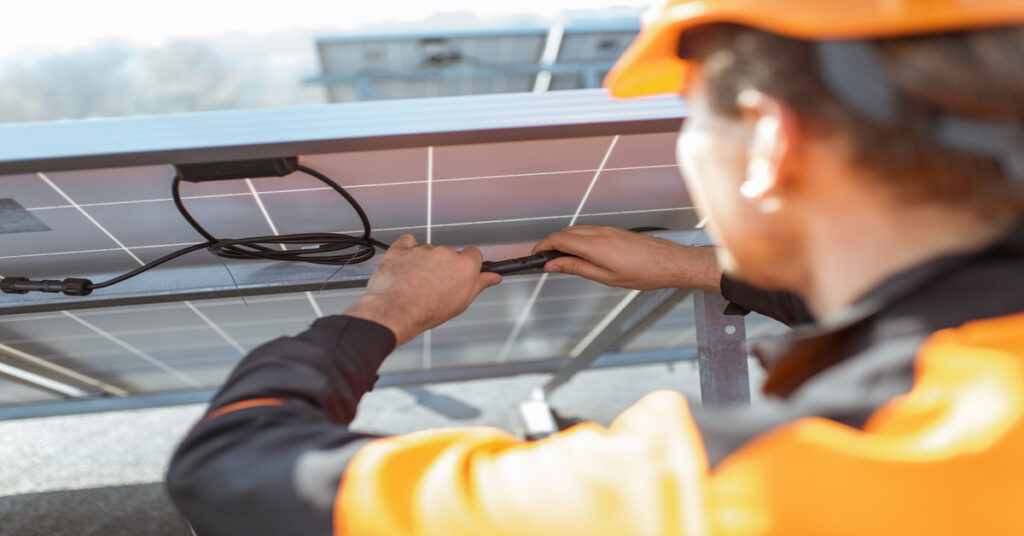 Solar Monitoring Systems Performance Data Access 1 A man wearing an orange uniform and a hat, plugging the cables in the back of a solar panel on a sunny day.