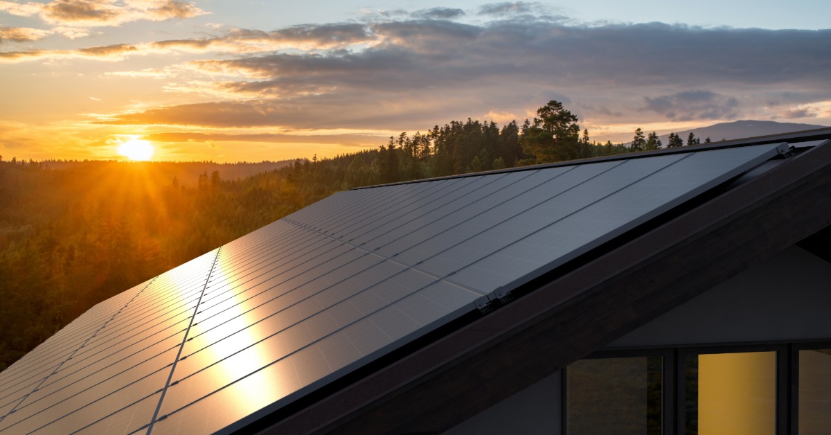 Microinverters vs. String Inverters for Home Solar Power 1 A close-up of a roof with multiple rows of solar panels surrounded by trees, with a sunset in the background.