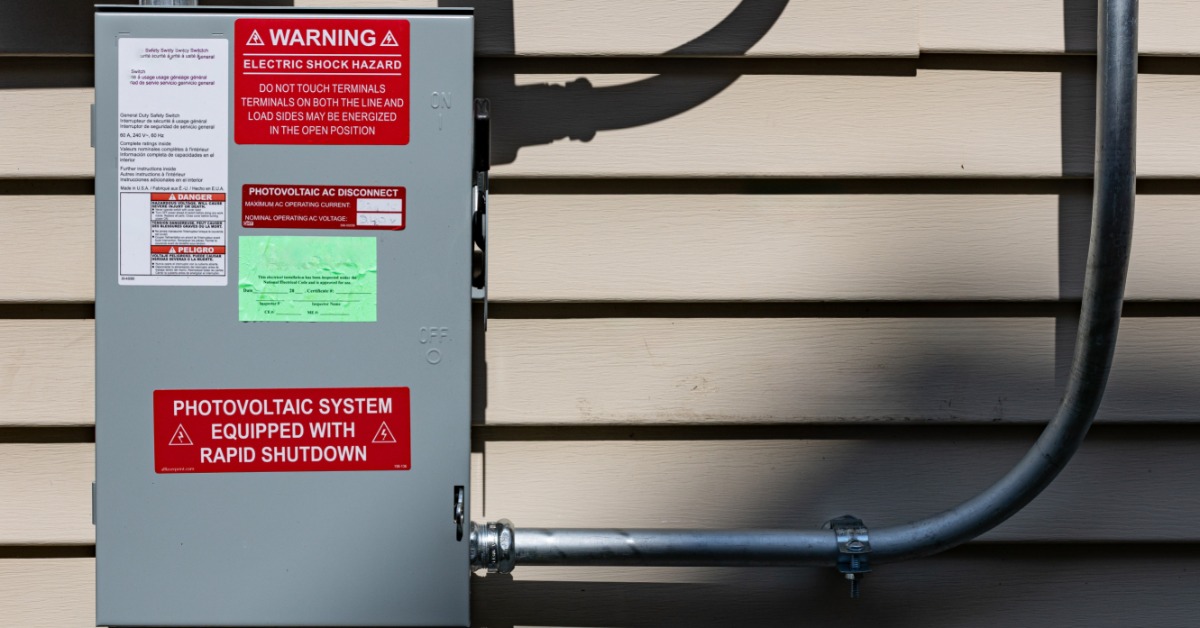 Microinverters vs. String Inverters for Home Solar Power 2 A large metal box with multiple warning labels on the front, attached to multiple tubes placed on a wall.