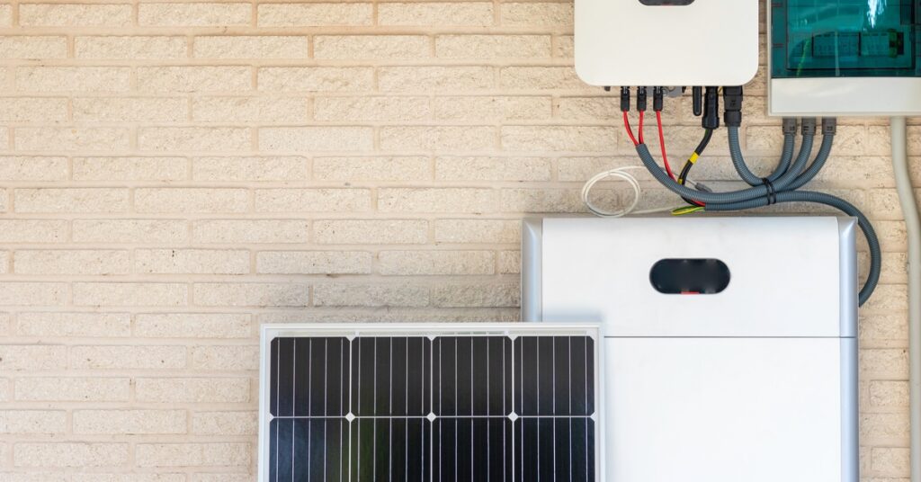 Lifespan of Critical Solar Electric System Components 1 A white, tall box placed on a wall under a smaller box with multiple red cables, behind a small solar panel.