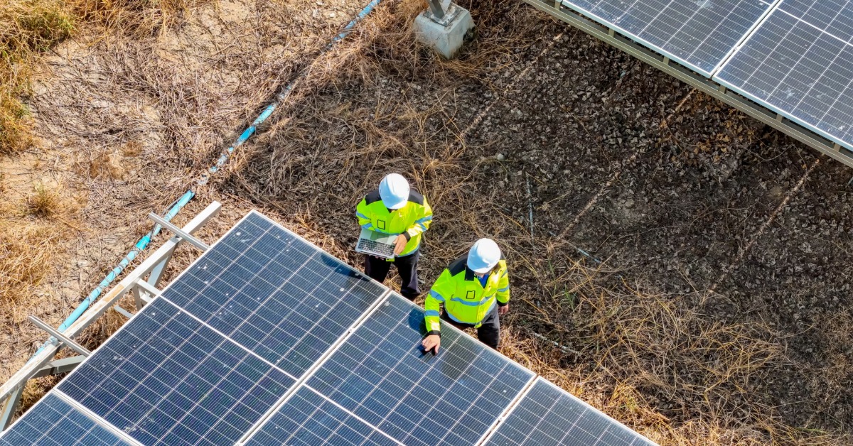 A Look at Ground-Mounted Residential Solar Power Systems 2 An aerial view of two workers wearing green jackets and helmets, walking between two rows of solar panels on the ground.