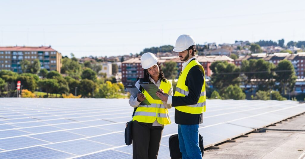 Three-Phase Power and Commercial Solar Integration 2 Two workers wearing yellow vests and white helmets, standing on a concrete roof with multiple rows of solar panels.