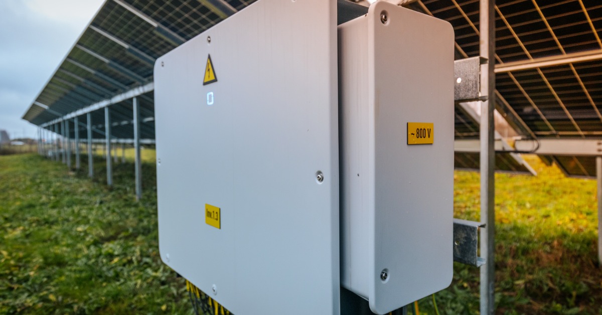Learn 1 A close-up of a large inverter box placed under multiple rows of commercial solar panels in an open grass field.