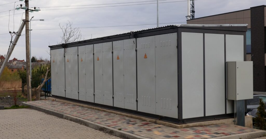 An Introduction to Tesla Megapack Utility Storage 1 A large white battery on a concrete slab with six pairs of doors, with electrical warning labels placed outdoors.