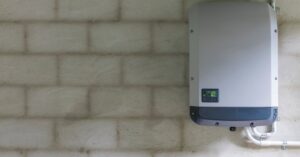 A grey rectangular power storage unit with a small screen in the bottom left, attached to a grey brick wall.