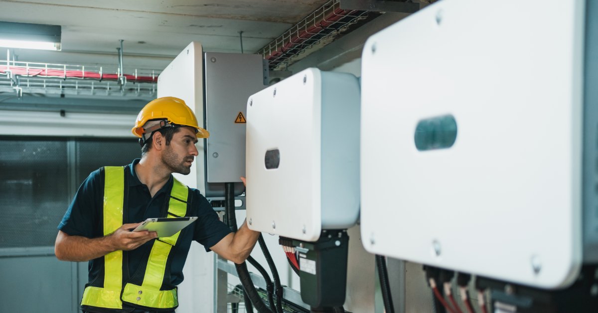 A Look at the Technical Specifications of the Powerwall 3 1 A man wearing a safety vest and a yellow helmet inspecting the side of a white battery between two other batteries.