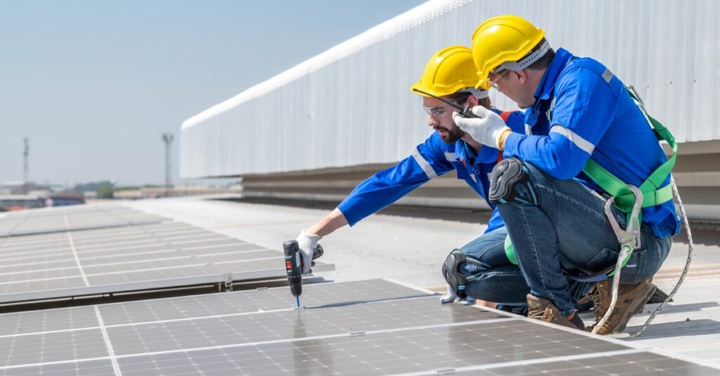 Scaling Solar for Manufacturing Facilities 2 Two workers wearing blue uniforms kneeling next to a solar panel while one uses an electric screwdriver on the panel.