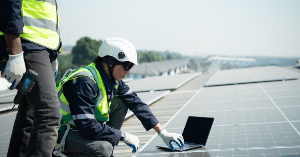 Scaling Solar for Manufacturing Facilities 1 A worker wearing a white helmet, kneeling next to a solar panel with a laptop on top, next to another worker.