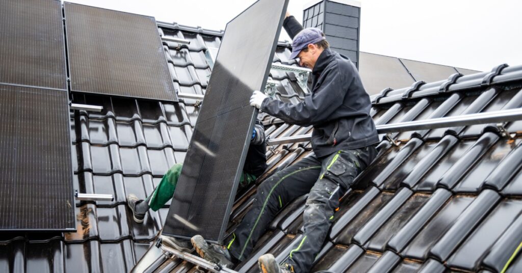Two workers placing a large solar panel on a very steep home roof with large shingles and mounting rails.