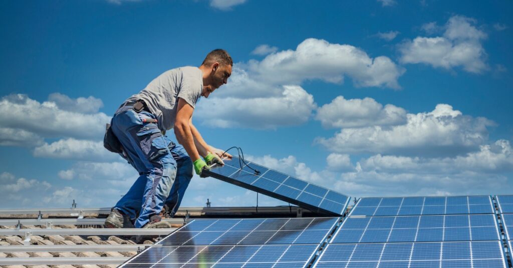 Two workers wearing grey shirts and safety gloves, placing a solar panel on the roof of a house with clay shingles.
