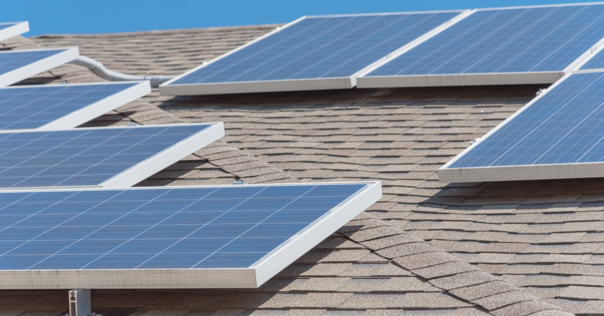 A close-up of multiple solar panels placed on a tilted roof with asphalt shingles, under the sun with a clear sky.